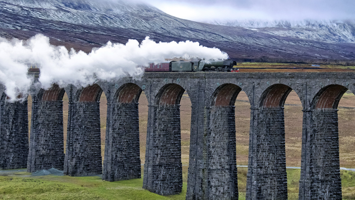 The West Yorkshireman service, hauled by a steam locomotive, travels on the Settle to Carlisle line over the Ribblehead Viaduct. The charter is pulling several brown carriages. The views span over the Yorkshire Dales National Park and the Pennines. 