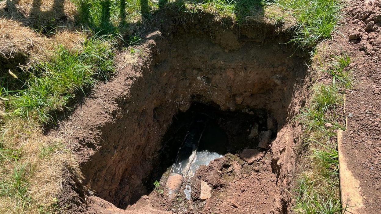 The image depicts a large hole dug into the ground, exposing a section of underground piping. Part of the piping remains exposed as brown sewage water makes its way through and overflows into part of the hole. The surrounding area is mostly grassy, with patches of dirt where the excavation has occurred.