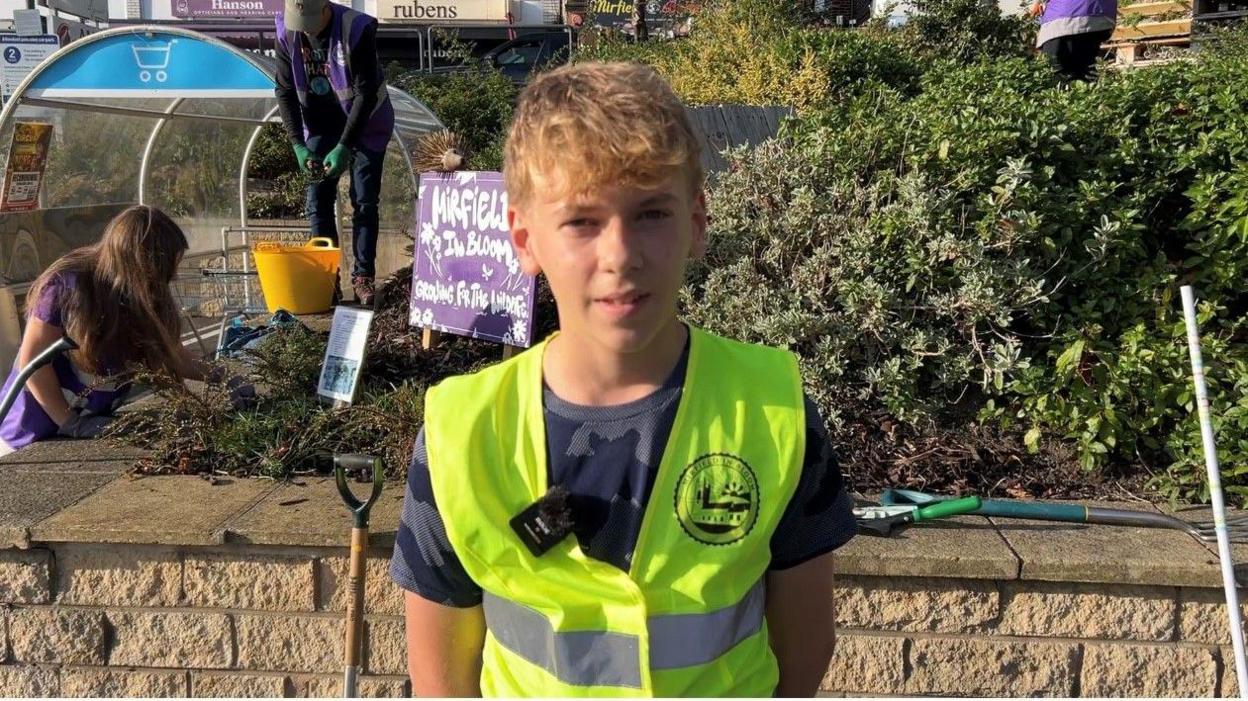 A teenage boy with blonde hair. He is wearing a high-vis jacket. Behind him is a wall with bushes and shrubs and volunteers doing some gardening.