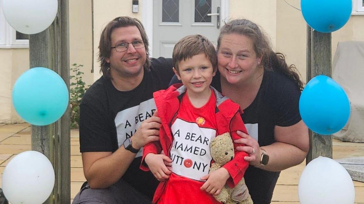 Riley with his parents Beth and Andy. They are stood under an arch which has been decorated with blue and white balloons. All are smiling and looking into the camera.