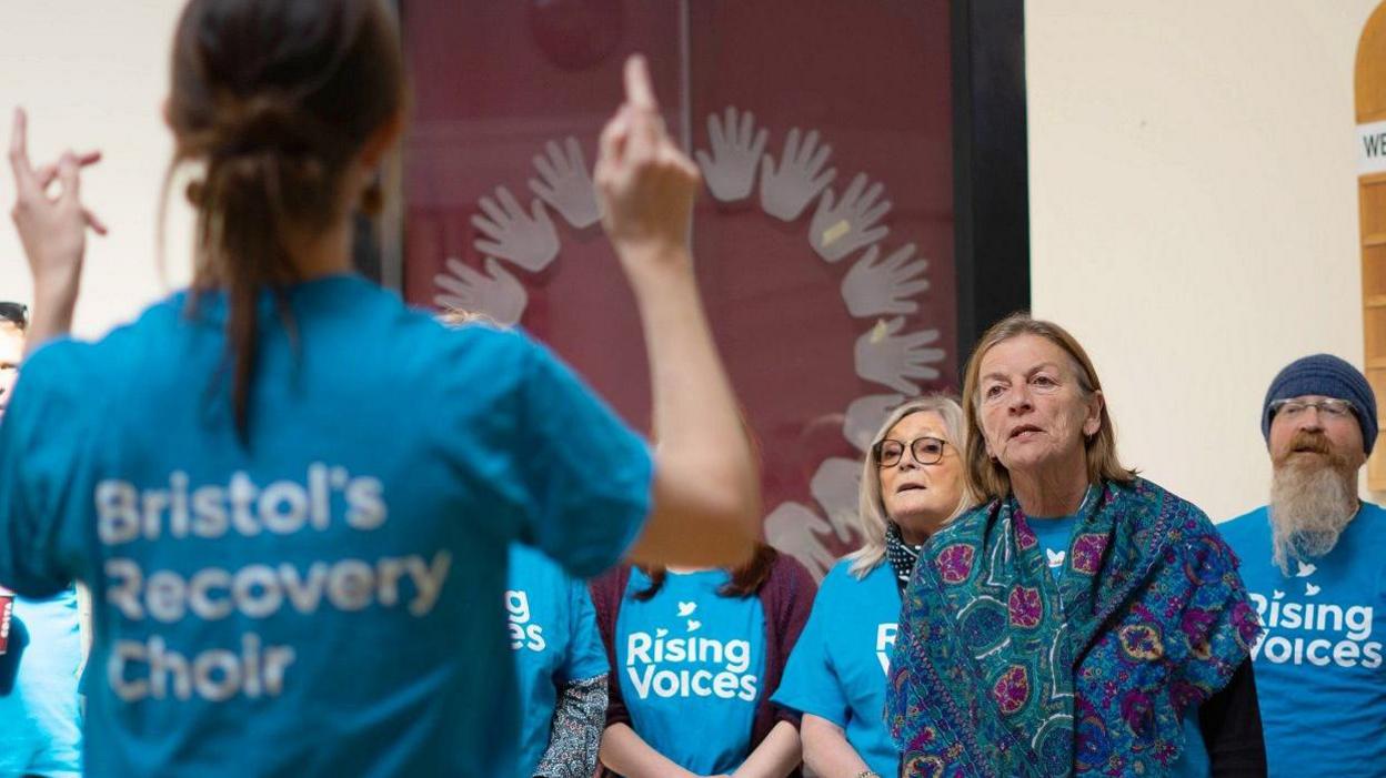 Members of the Rising Voices choir in Bristol face their conductor as they sing. They are all wearing blue T-shirts with the name of the choir written on them in white lettering.