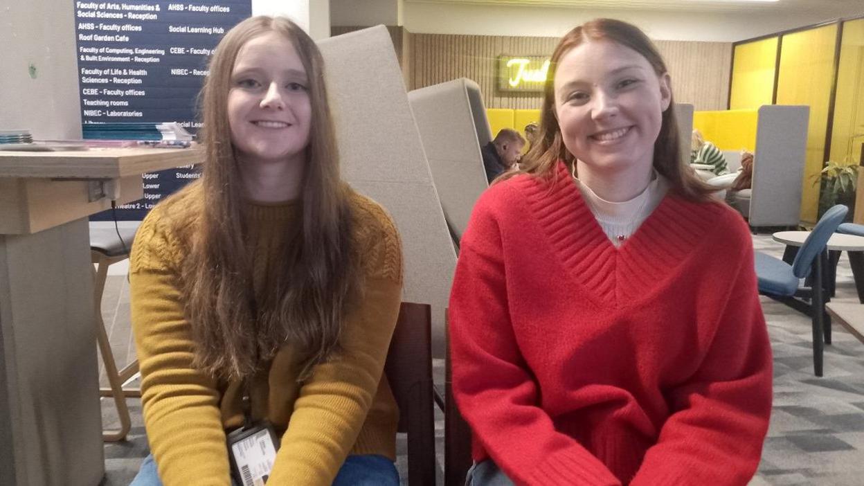 Carenza Gamble sitting down with long, light brown hair, wearing a mustard coloured jumper beside Lucy Belle Rodgers who has shoulder length brown hair and is wearing a red jumper