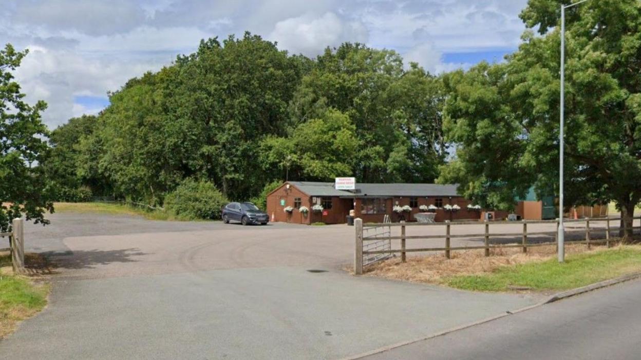 A long brown building, set back from the road, with a grey roof. There are large trees behind the building and a large concreted area for cars.