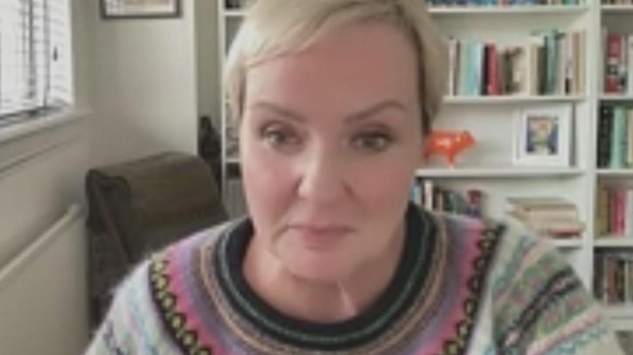 Zoom call grab of Susan Browne. She is wearing a woollen jumper with a Scandi pattern. She has short blonde hair. Behind her is a white book shelf containing books, ornaments and photos. 