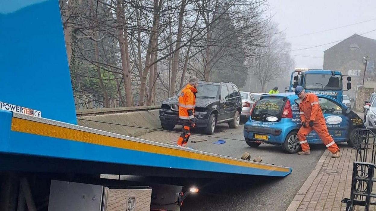 Two workmen clad in orange overall haul a blue car onto a towing vehicle. It's a cloudy day and leafless trees are in the background, along with a number of other parked cars.