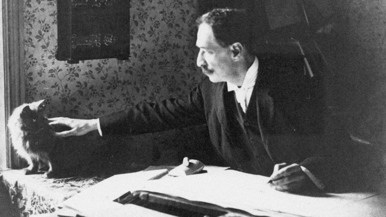 A black and white photo of Louis Wain from the 1890s sat at a desk with a pen and drawing book whilst stroking a cat beside him