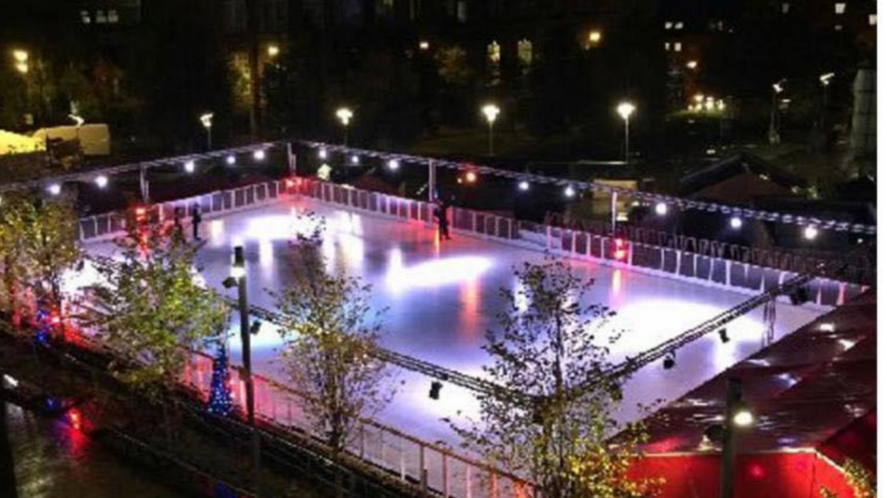 A mostly empty outdoor ice rink at night, rectangle shaped with spotlights surrounding it. Trees are dotted on the outside of it. 