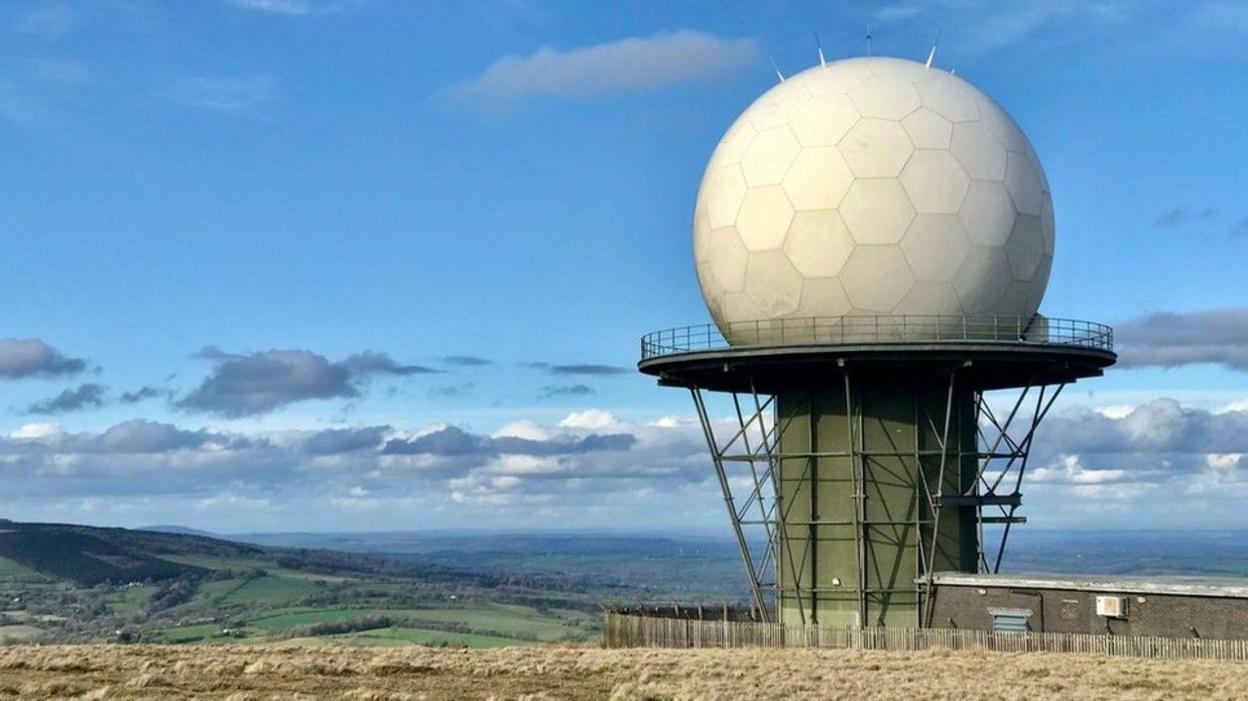 In the foreground there is a dome, that looks like a golf ball, atop a platform and tower. Countryside is in distance and much of the image features blue sky or clouds.