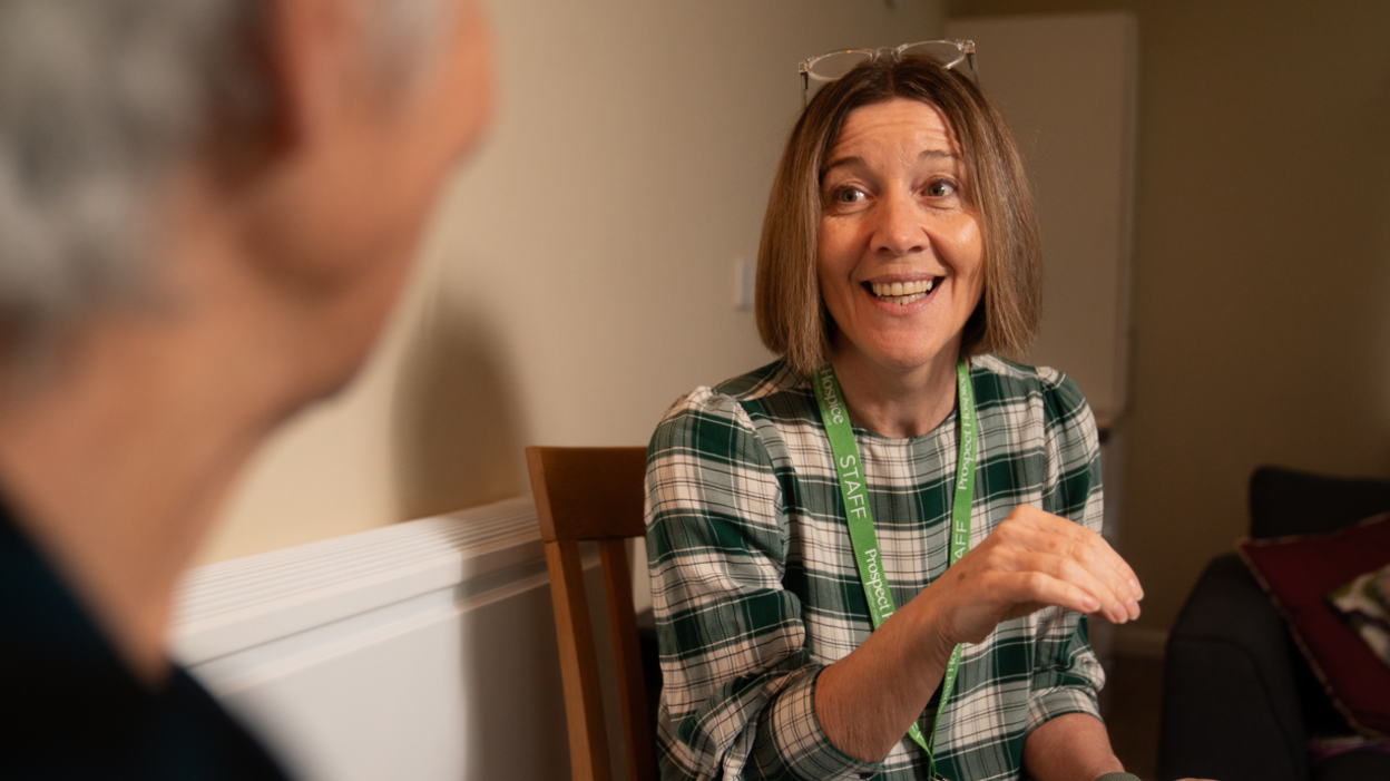 A woman talking to another woman while smiling. The woman who is talking has brown hair in a long bob haircut and she is wearing a green and white checked top. She has glasses balanced on her head and she is wearing a green lanyard that says 'staff' on it. The other woman is to the left of the picture and is out of focus.