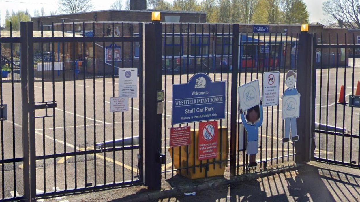 Outside of Westfield Infant School - a gate around the school is at the front of the photograph with signs on, and the playground and building is in the background. 