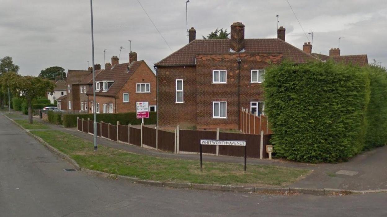 One end of a street of two-storey brick houses with white-framed windows, enclosed by low wooden fences. There is a white street sign saying "Whitworth Avenue" in the centre of the shot. There is a tall hedge to the right and a road with a grass verge sweeps round to the right.