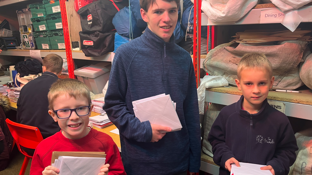 Three young boys standing together in a sorting office holding piles of Christmas cards in envelopes. On the left, a boy with fair hair and black-rimmed glasses wearing a red jumper. In the middle, an older boy who is much taller in a blue fleece. On the right, a shorter and young boy, wearing a navy-blue or purple fleece.