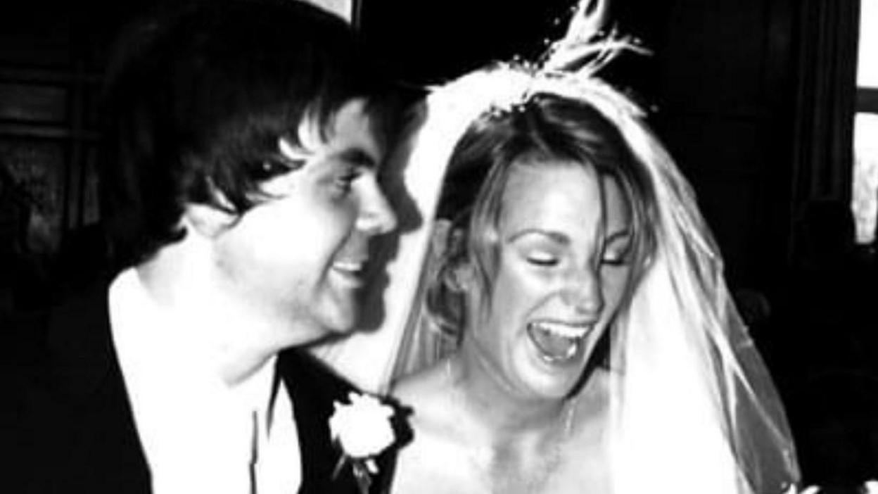 A black and white photo of Geraint and Deb's wedding day. He is in a suit with a buttonhole and she is wearing a veil and laughing.  