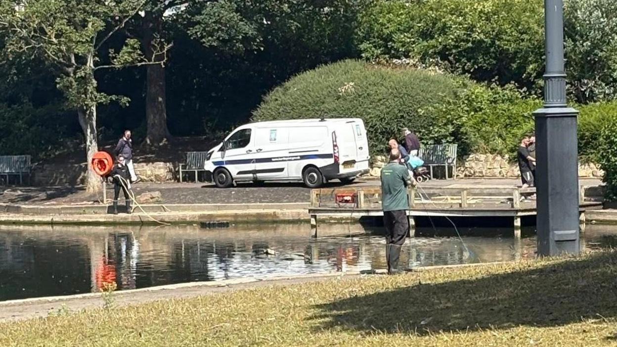 A white van is parked at the side of a lake with lots of people holding pipes running into the water.