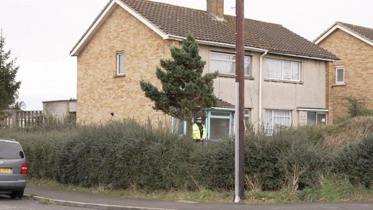 A police officer stands outside a semi-detached house with their hands clasped in front of them. They are wearing full uniform, including a hat and a high-vis coat. The house is built of light brick and has a pale rendering on the front, and there are are a number of large bushes in the front garden. A grey car parked on the street is partially visible. 