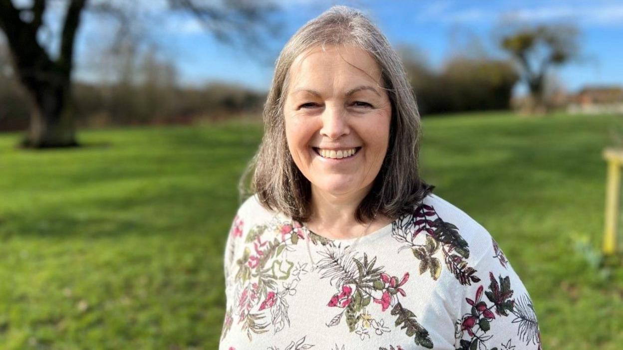 A woman with grey hair and wearing a white top including partially red and green flower patterns is smiling at the camera. A field behind her and trees are blurred in the background.