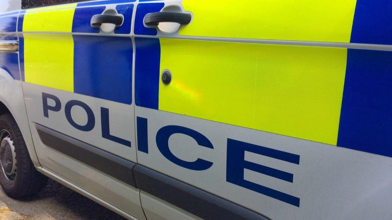 The left hand side of a police van with the word police marked in blue lettering below a yellow and blue fluro strip.