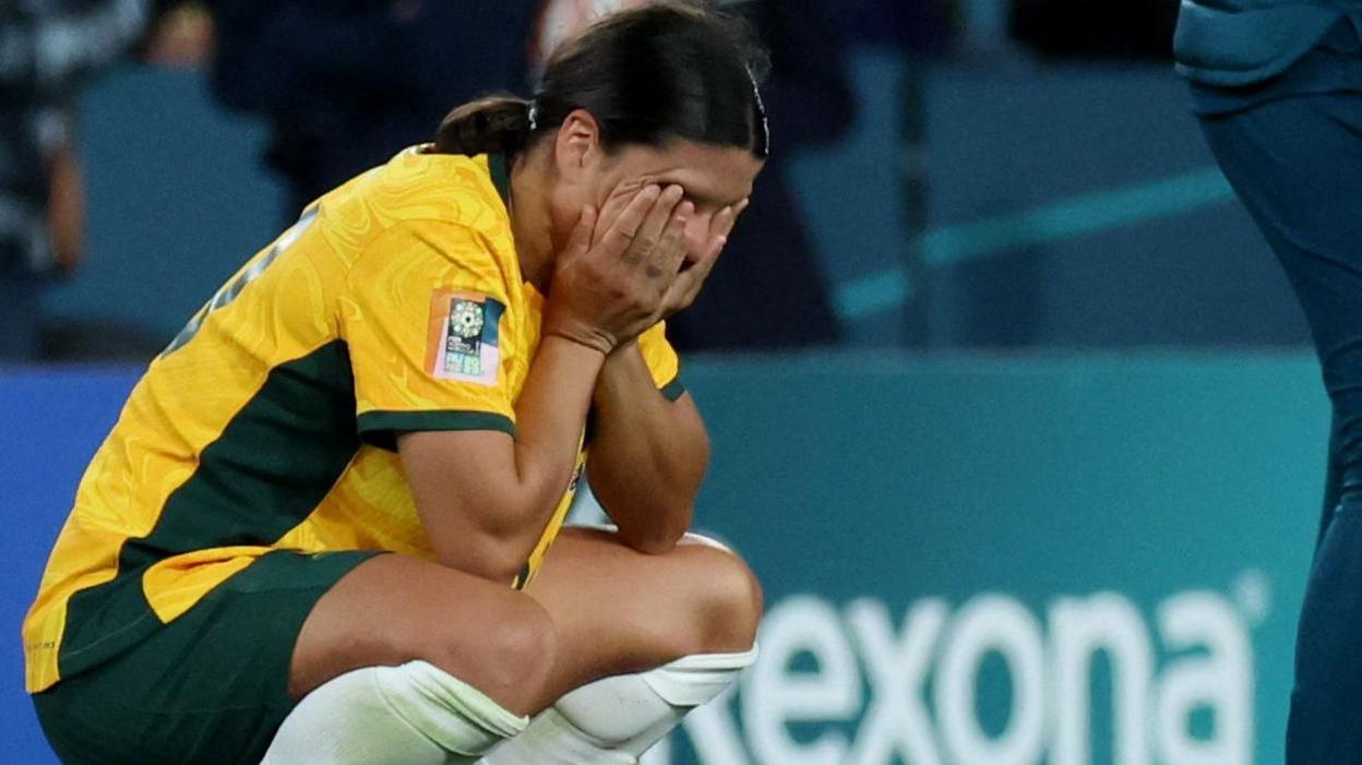 Sam Kerr crouching down looking disappointed on a football pitch, wearing Australian national strip which is yellow and green.