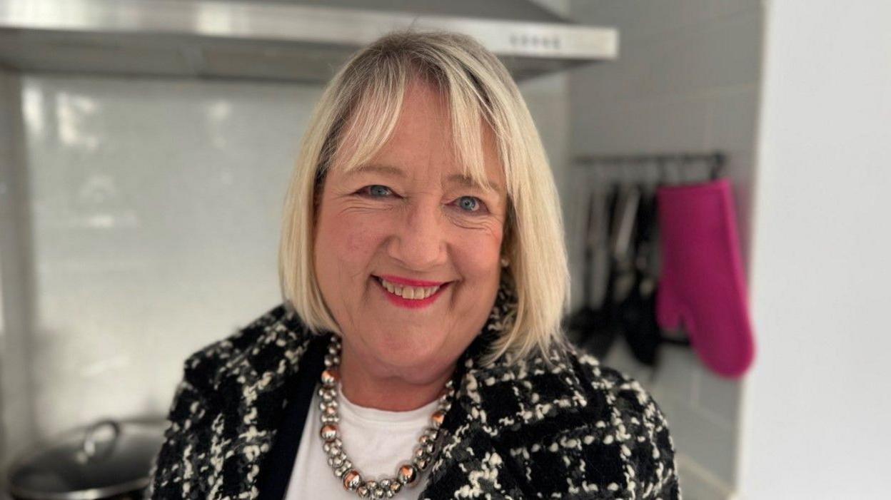 Jane Jones, with blonde hair, is smiling at the camera and wearing a black and white coat, white top and necklace. Kitchen equipment is blurred behind her.