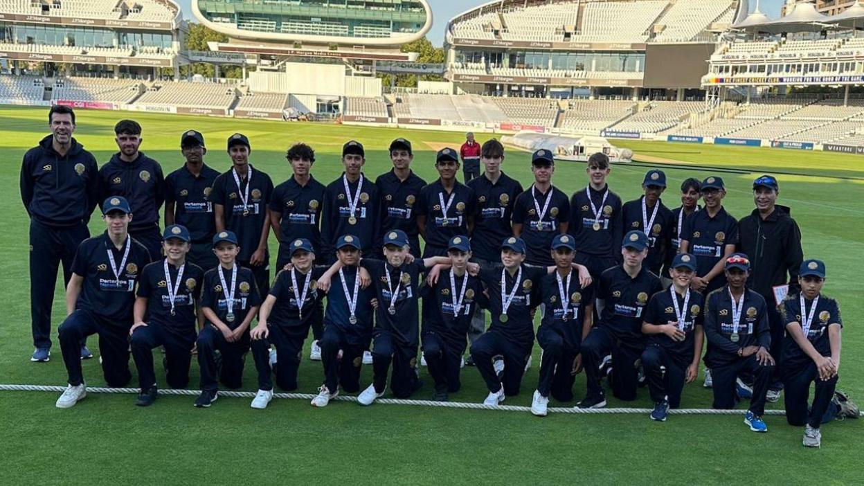 Two youth cricket squads, dressed in navy t-shirts, trousers and caps, lined up for a team photo on Lord's ground. There are 12 members wearing medals and three adult coaches.