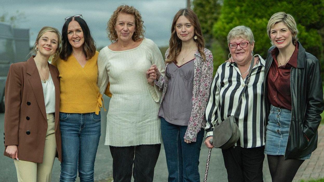 Three female cast members stand smiling with their real-life counterparts. They are on a set that looks like a residential street.