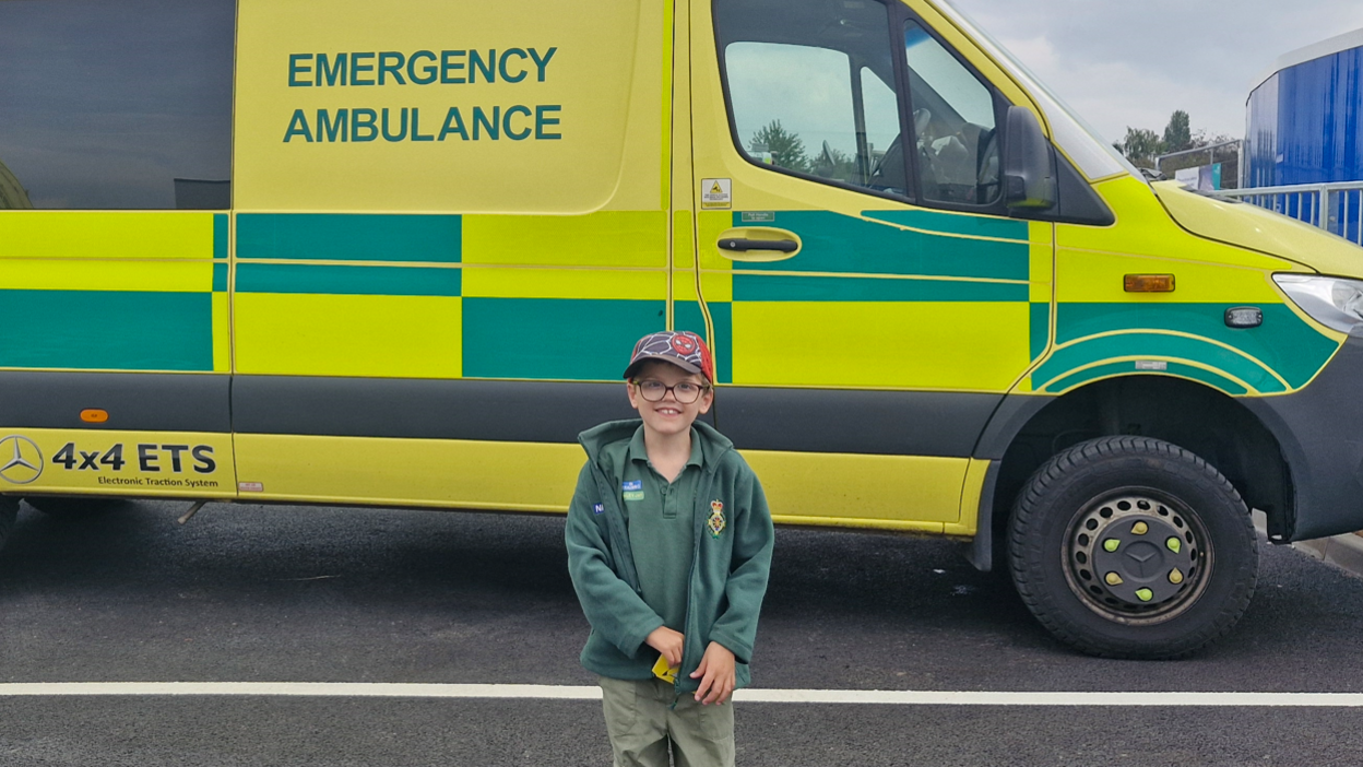 A boy with glasses, a green tshirt and fleece and a red cap standing in front of a green and yellow chequed ambulance