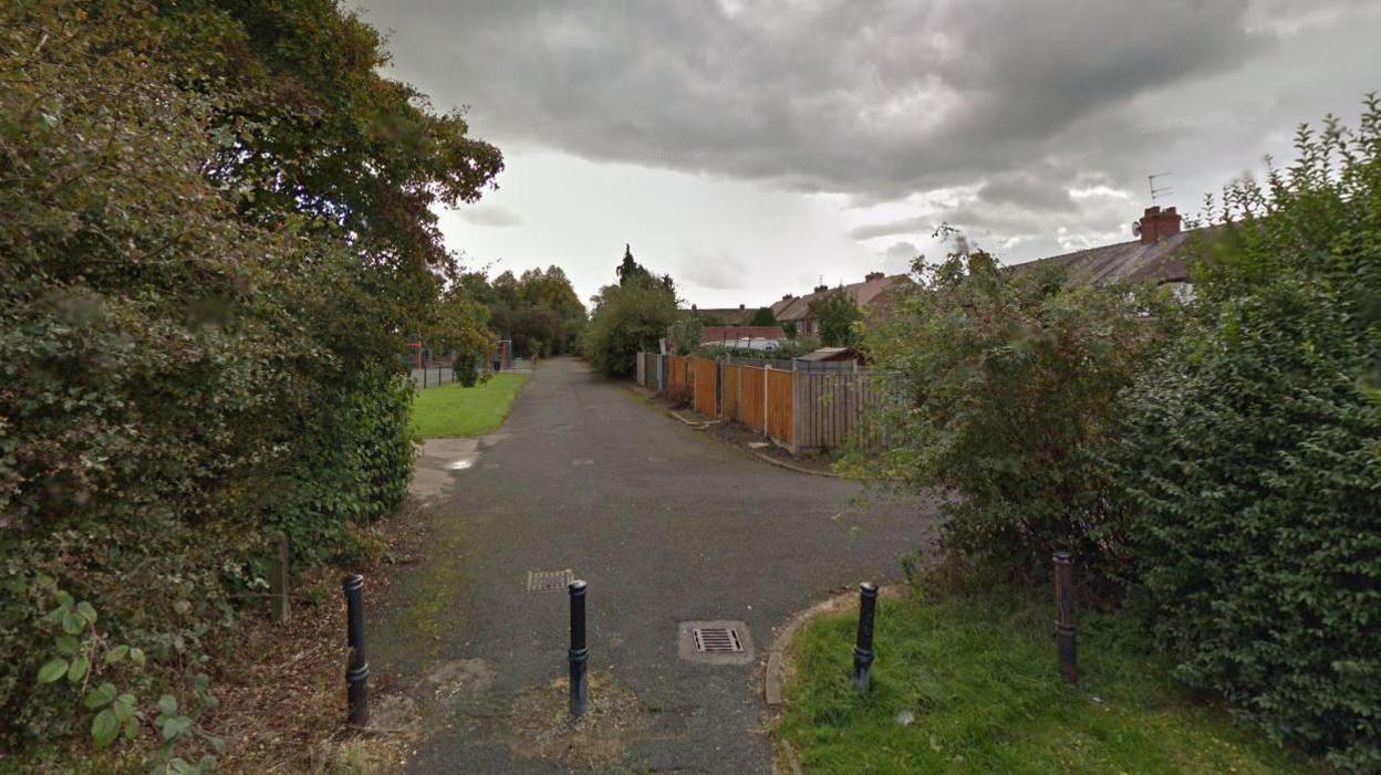 An entrance to Baguley Lane, in Sale which runs at the back of houses. Pictured is four black posts on a path, with the path stretching in the background next to a park. 