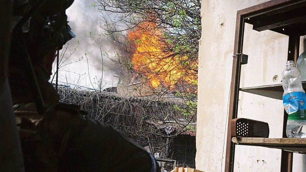 A unformed Ukrainian soldier stands outside, watching a building burn