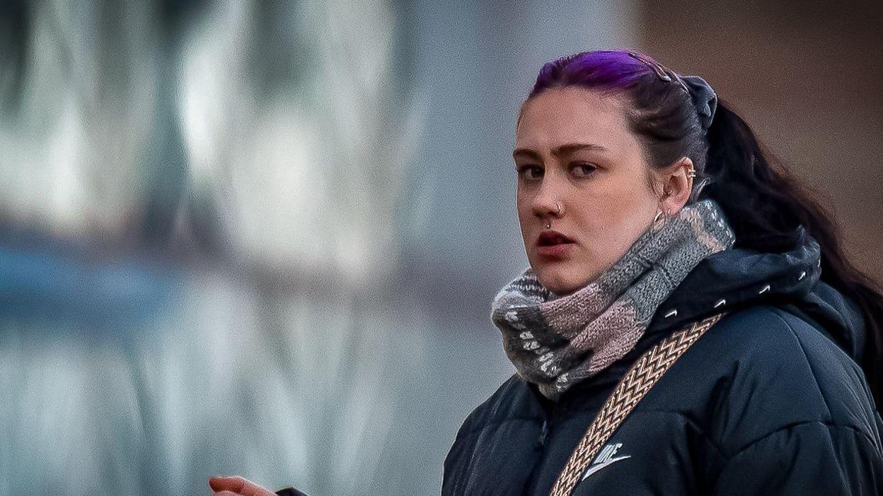 Rhiannon Evans arriving outside court. She has pulled back long black hair with a purple streak around her top part and is wearing a puffy black winter coat.