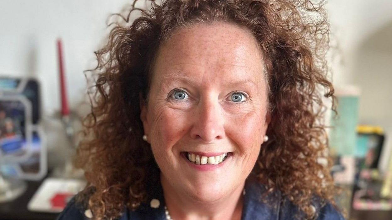 A woman smiling with curly brown shoulder-length hair, blue eyes wearing pearl earrings and necklace, a dark blue shirt with a blurred background 