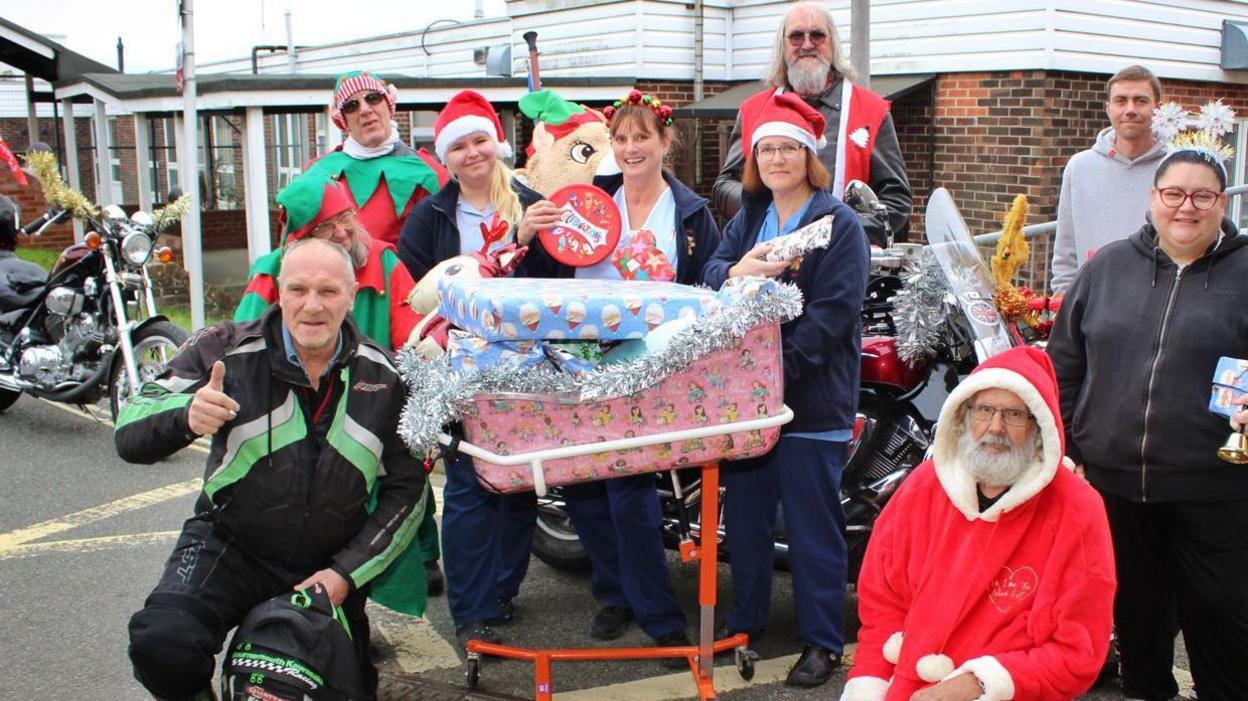 Some of the bikers posing for the camera with hospital staff. Some of them are dressed up as Santas. The hospital staff is around a cot filled with presents. One of the nurses is holding a box of chocolates. One biker is dressed in regular clothes and giving thumbs up. There is a bike with tinsel in the background.