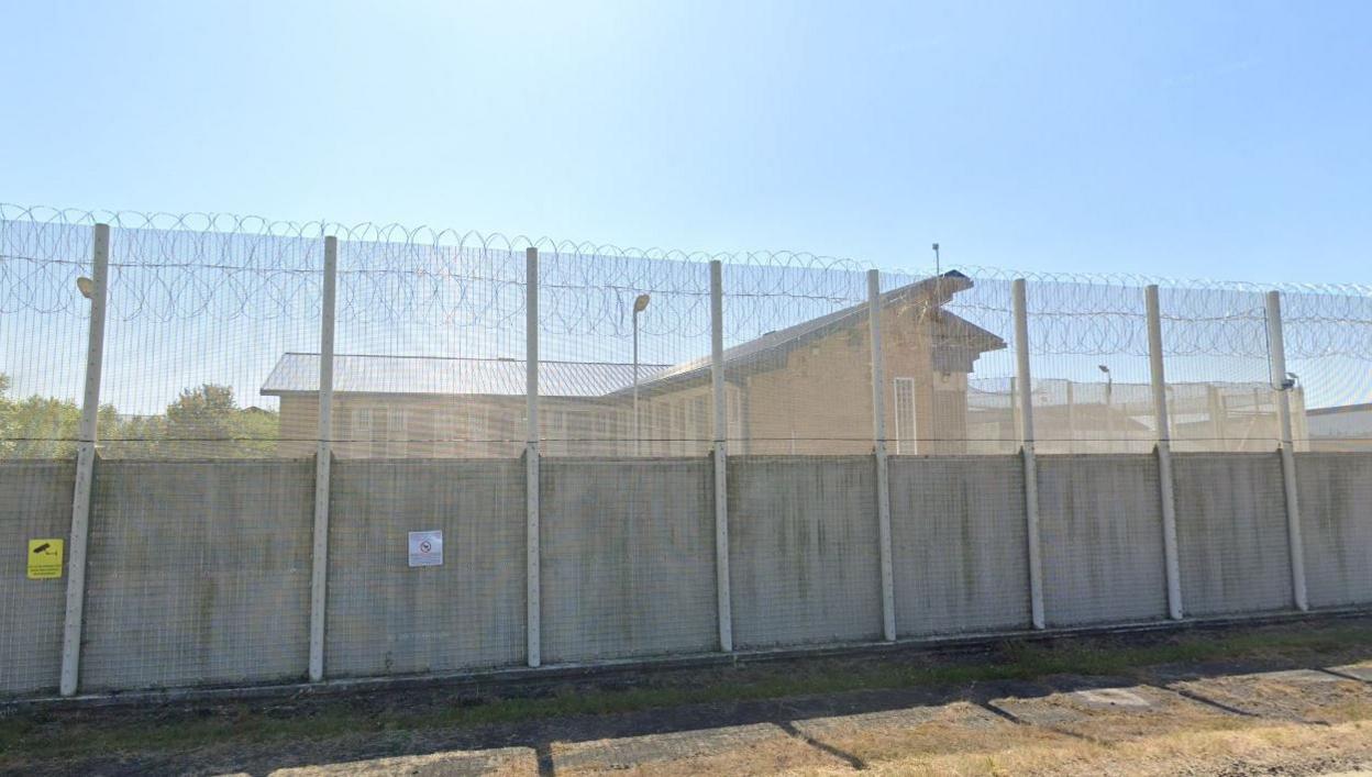 Streetview of a large prison fence with buildings beyond