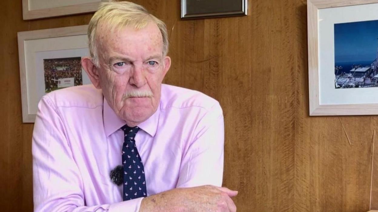 David Harris, who has grey hair and moustache, is wearing a pink shirt and tie, and sitting down in front of a wood-panelled wall with framed pictures on it