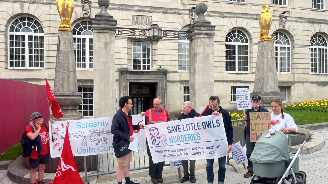 Seven campaigners hold banners and flags outside a pale brick building. Gold owls stand on pillars. Plaques read "Save Little Owls Nurseries."