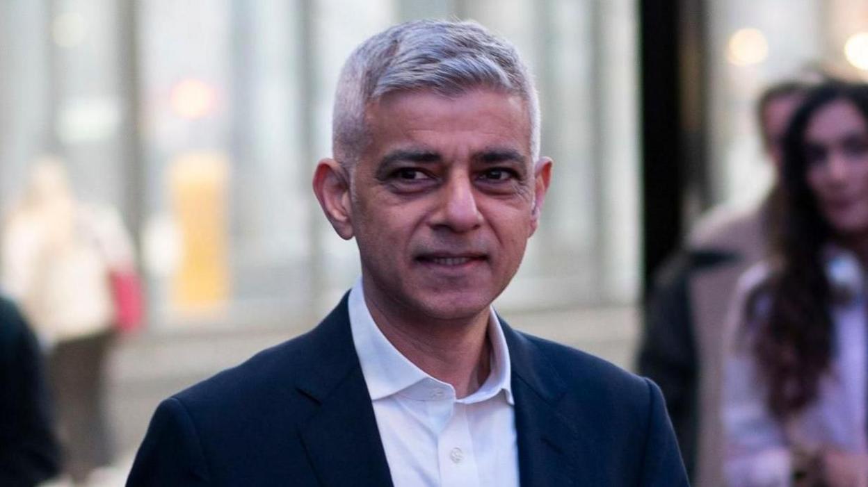 London mayor Sadiq Khan is wearing a dark suit jacket and a white shirt. There are people standing to the side taking photos of him.
