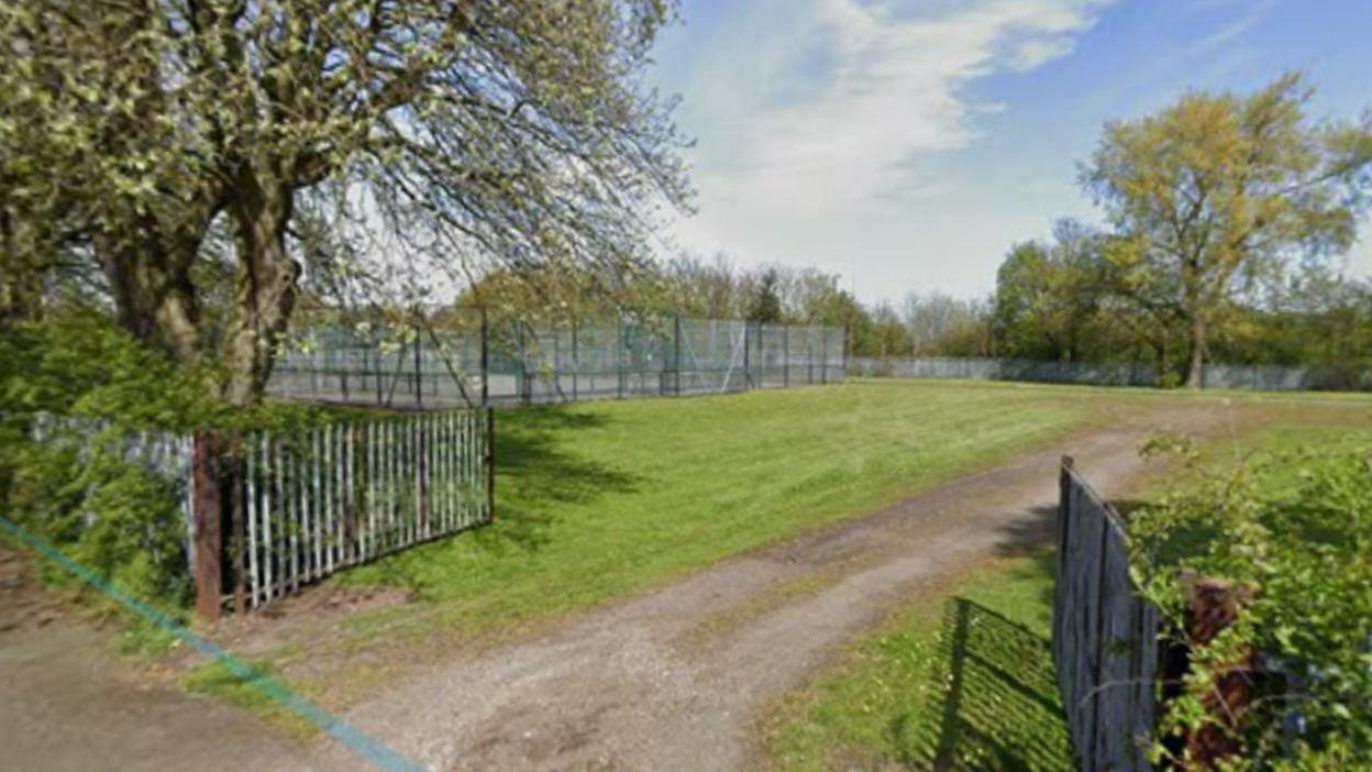 View of a grassed area fronted by two gates with vertical metal bars, which are open, and a gravel track leading up between them. Behind the fenced-off grassed area there are a number of trees and shrubs.