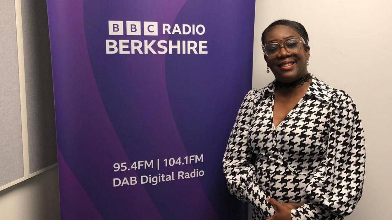 A woman with glasses and wearing a smart black and white patterned dress stands in front of a purple sign for BBC Radio Berkshire.