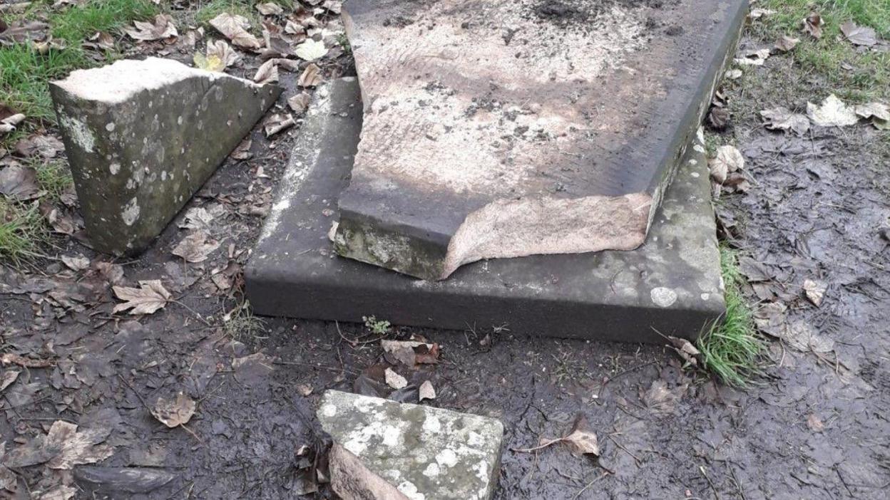 A gravestone on a patch of muddy ground is seen broken into a number of pieces.
