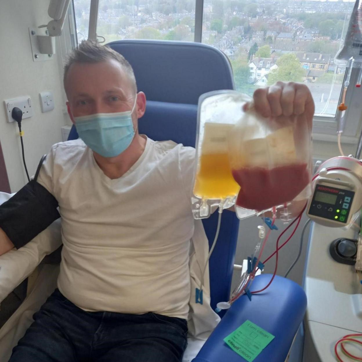 A man sitting in a blue hospital chair in front of a window while holding two blood bags, one containing a dark-red substance and the other an amber liquid. He has short light hair and is wearing a blue facemask and a white T-shirt. A black band is strapped around one arm. There is medical equipment next to him.