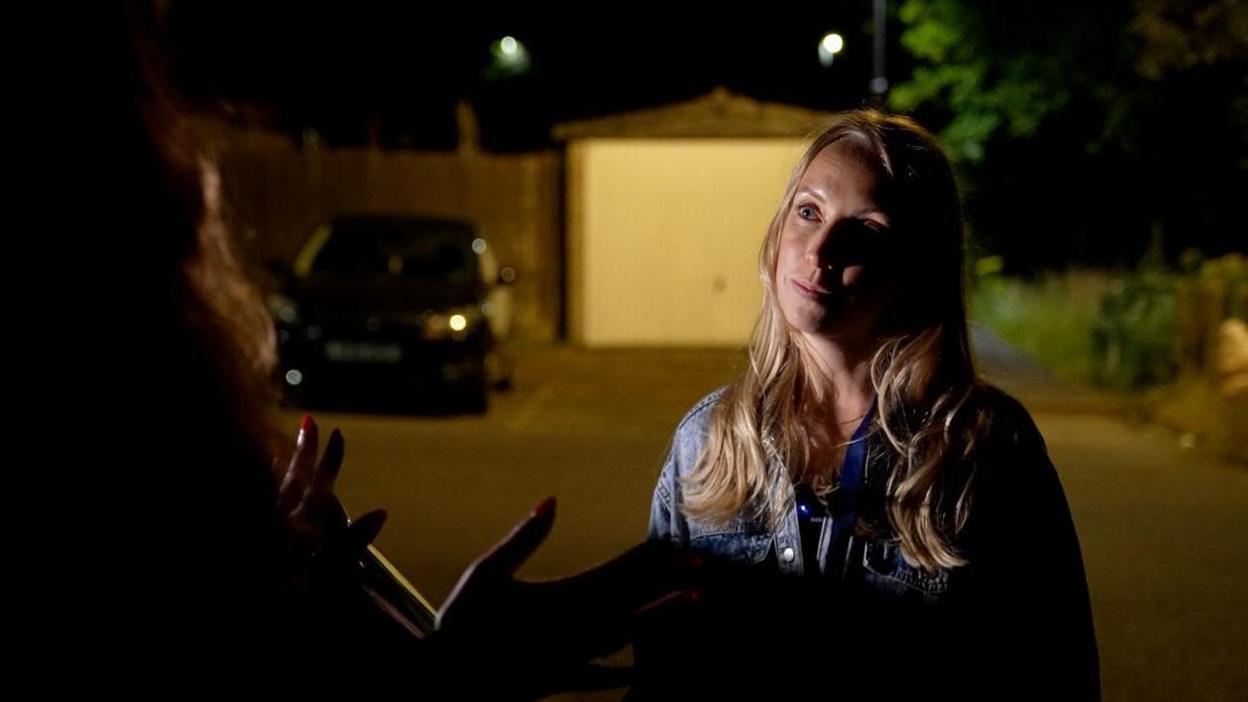 Jo Ritchie stood in a car park at night. There is a garage and bushes behind her. It is night time and her face is lit with a light from a camera. Behind her is dark. She has long blonde hair and is wearing a denim jacket.