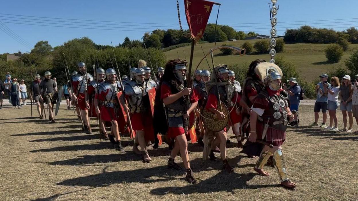 A group of people dressed as Roman soldiers march in formation through a field, watched by onlookers. They are all wearing ornate red costume, and full armour. 