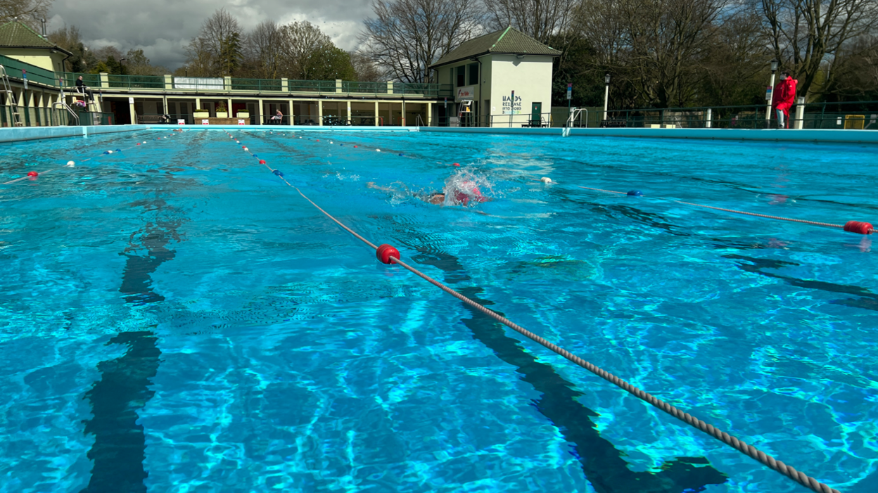 Peterborough Lido, which has separate swimming lanes. A single swimmer is in the pool. The sky is cloudy. The water is bright blue and there is a lifeguard dressed in red at the side of the outdoor pool.