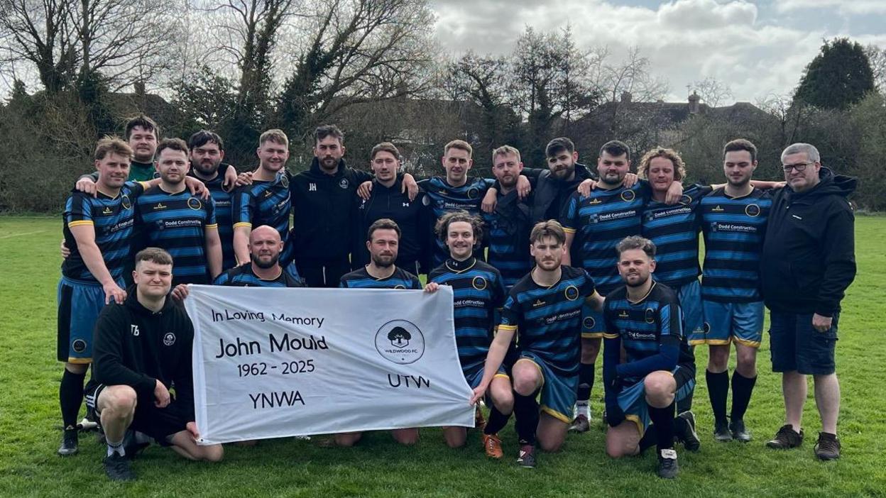 A team of footballers wearing blue and black stripes kits, stand on a football pitch and hold a white flag saying "In loving memory, John Mould, 1962-2025, YNWA". 