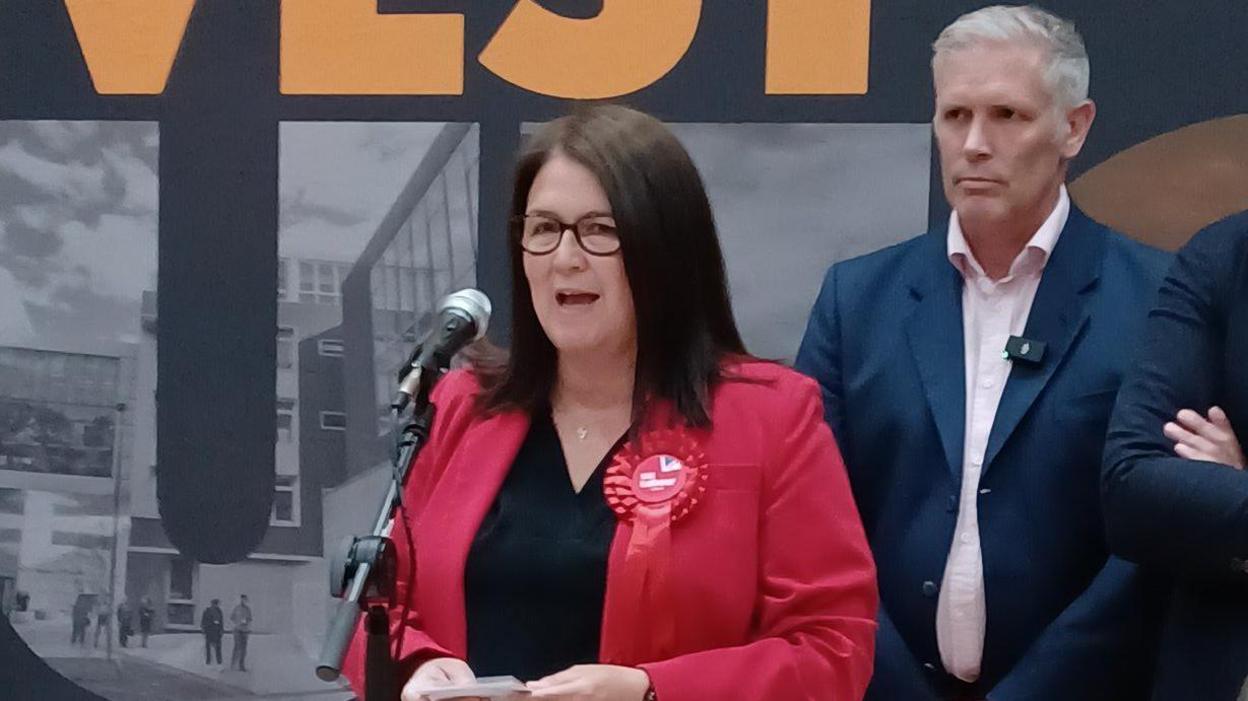 Rachel Hopkins in a red jacket with a red rosette attached to its lapel speaks into a microphone while holding a small set of notes. She has dark hair and wears glasses, and a man stands behind her.