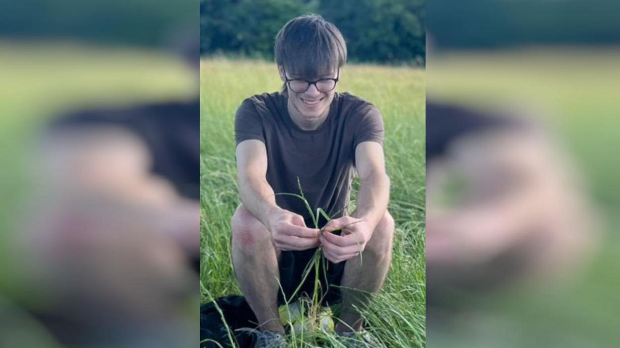 A man sits in a field playing with long pieces of grass.