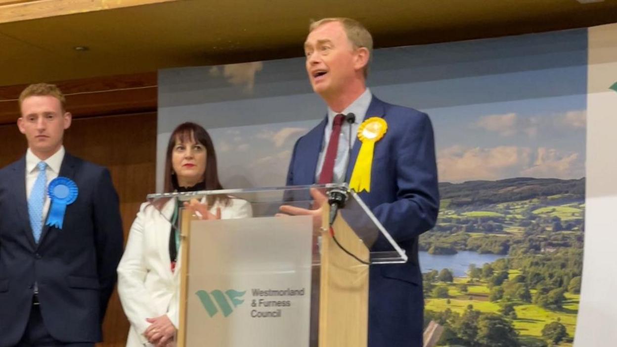 Tim Farron, wearing a suit and a yellow rosette, gives a speech behind a podium