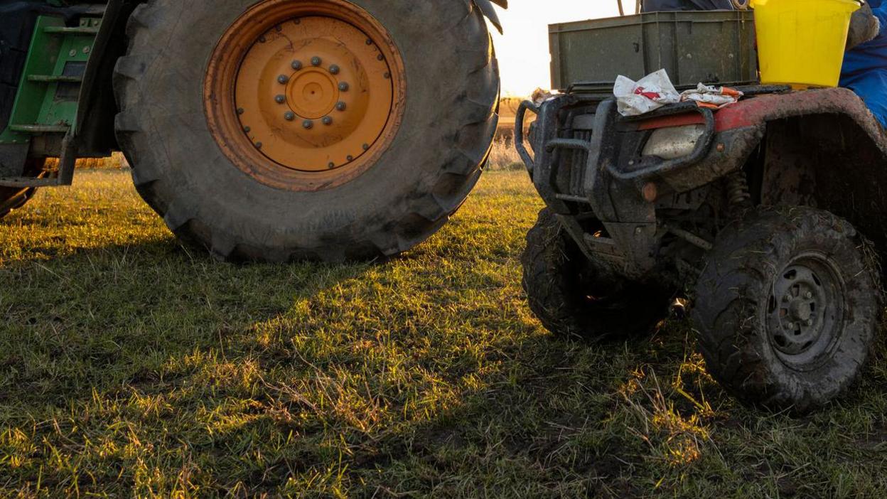 A close-up shot of a generic picture of farming machinery. It shows a red quad bike on grass with hay and mud on it. There are two people sitting on it, who are partly shown and are wearing blue. One is holding a yellow bucket. There is a large green tractor next to it, but you can just see the wheel.