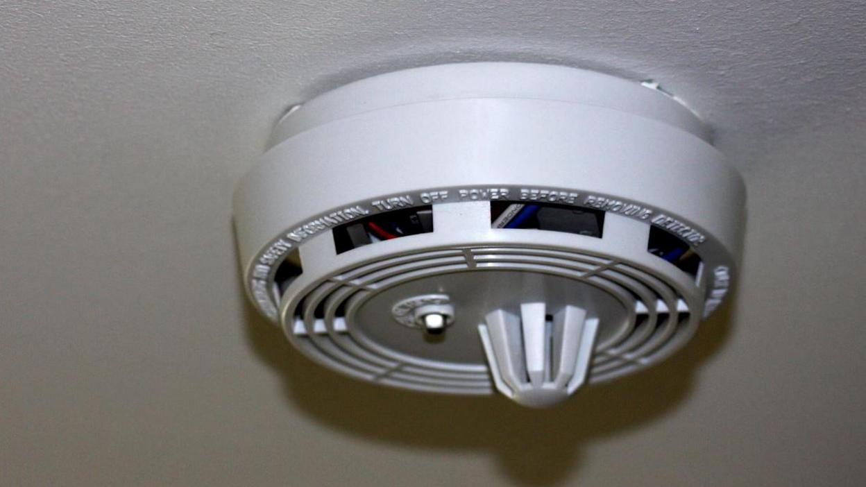 A close-up of a domestic smoke alarm on a white ceiling in a lit room with instruction of use on it