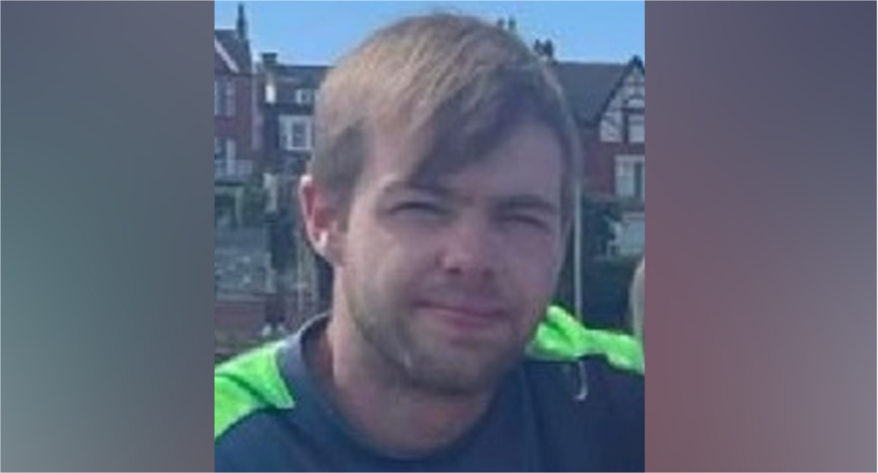 Man with brown hair and long fringe - he is wearing a grey t-shirt with neon lapels  - sitting in front of houses 