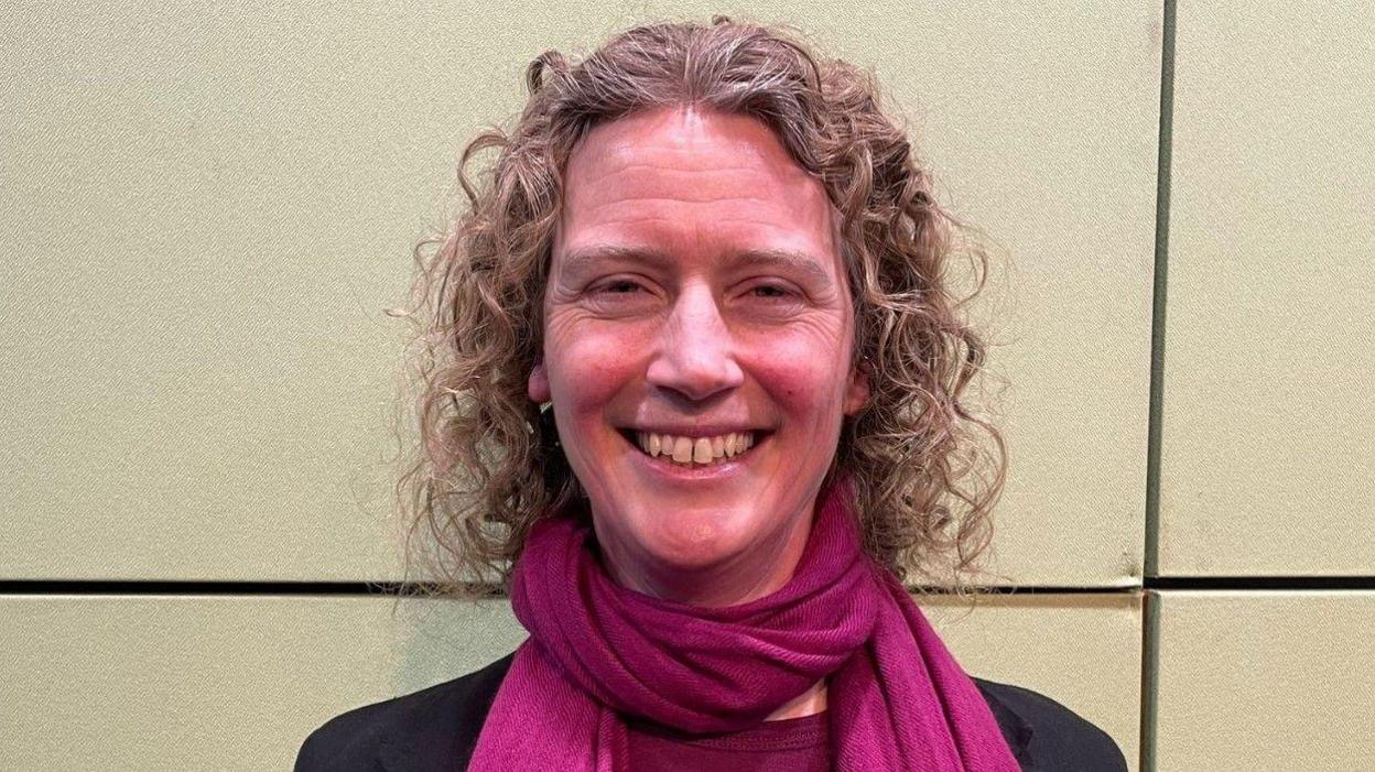 Becky Frall with curly hair is smiling and wearing a purple scarf. She is smiling in front of a yellow background.